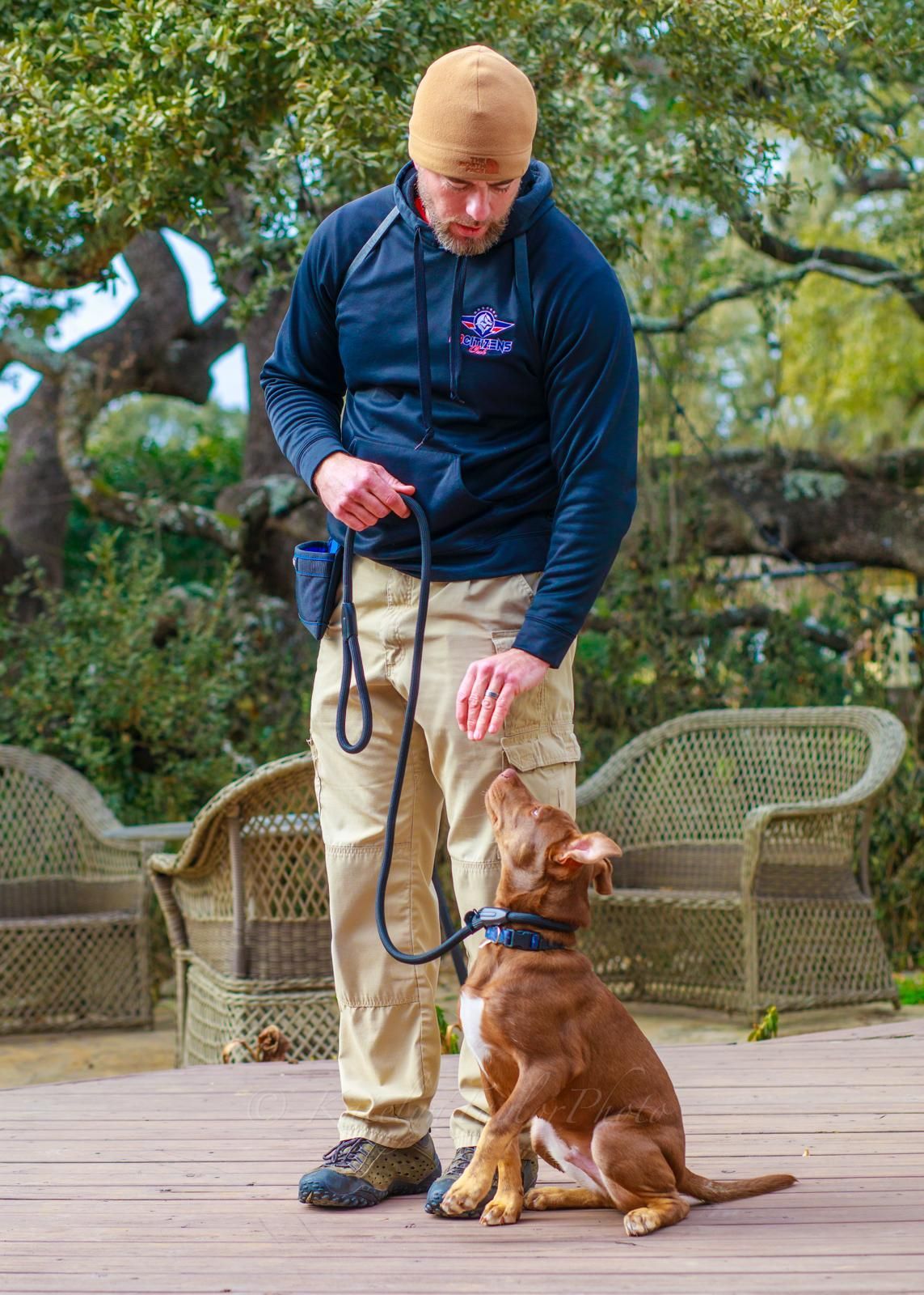 Man in black jacket training brown dog on a deck. The dog is looking up with leash attached.