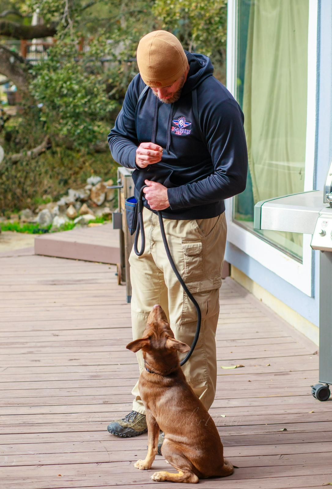 Man in khaki pants and black hoodie holding a leash, training a brown dog on a wooden deck.