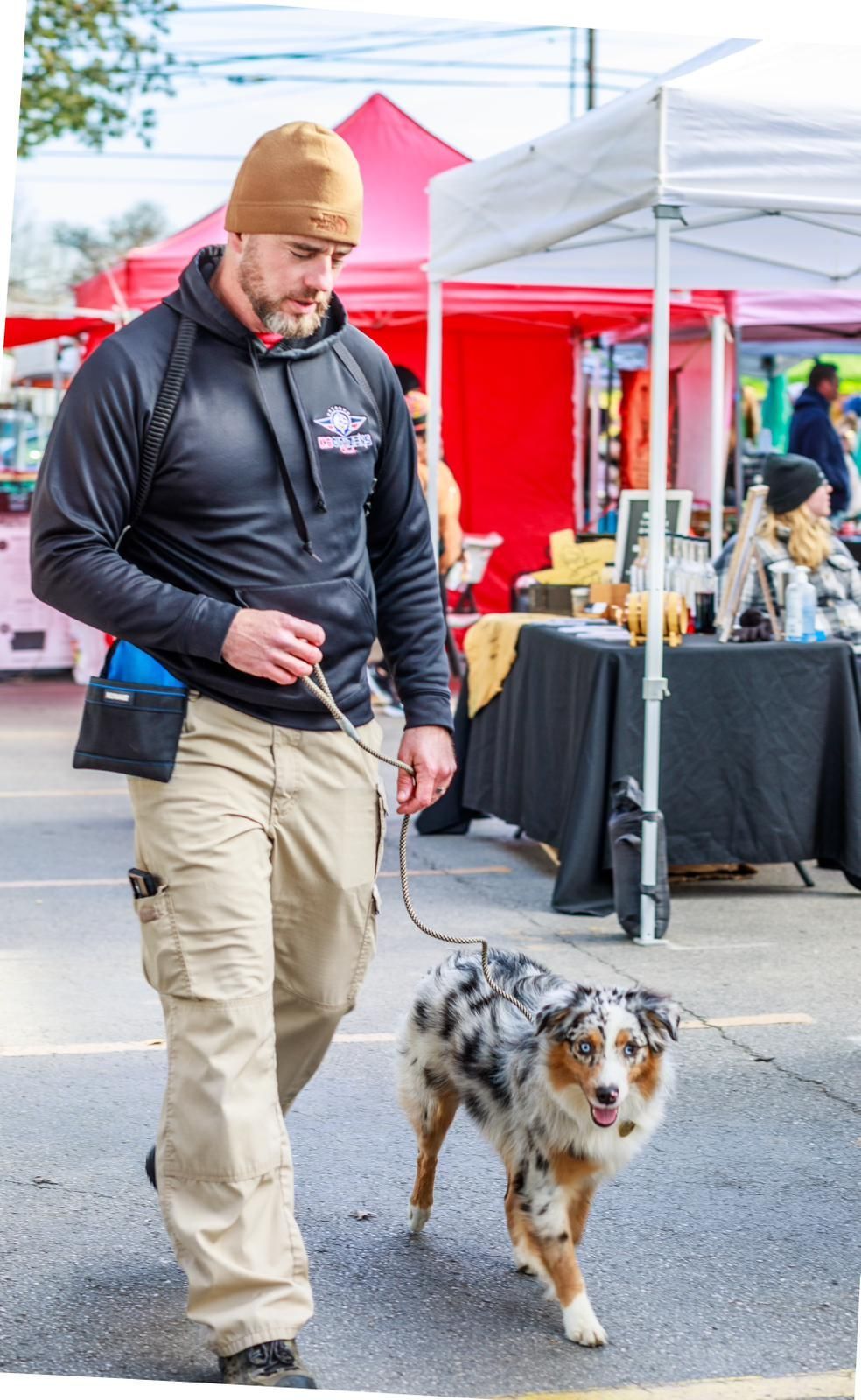 Man walks an Australian Shepherd at an outdoor market. Man wears a cap, black hoodie, and khaki pants. The dog has blue merle fur.