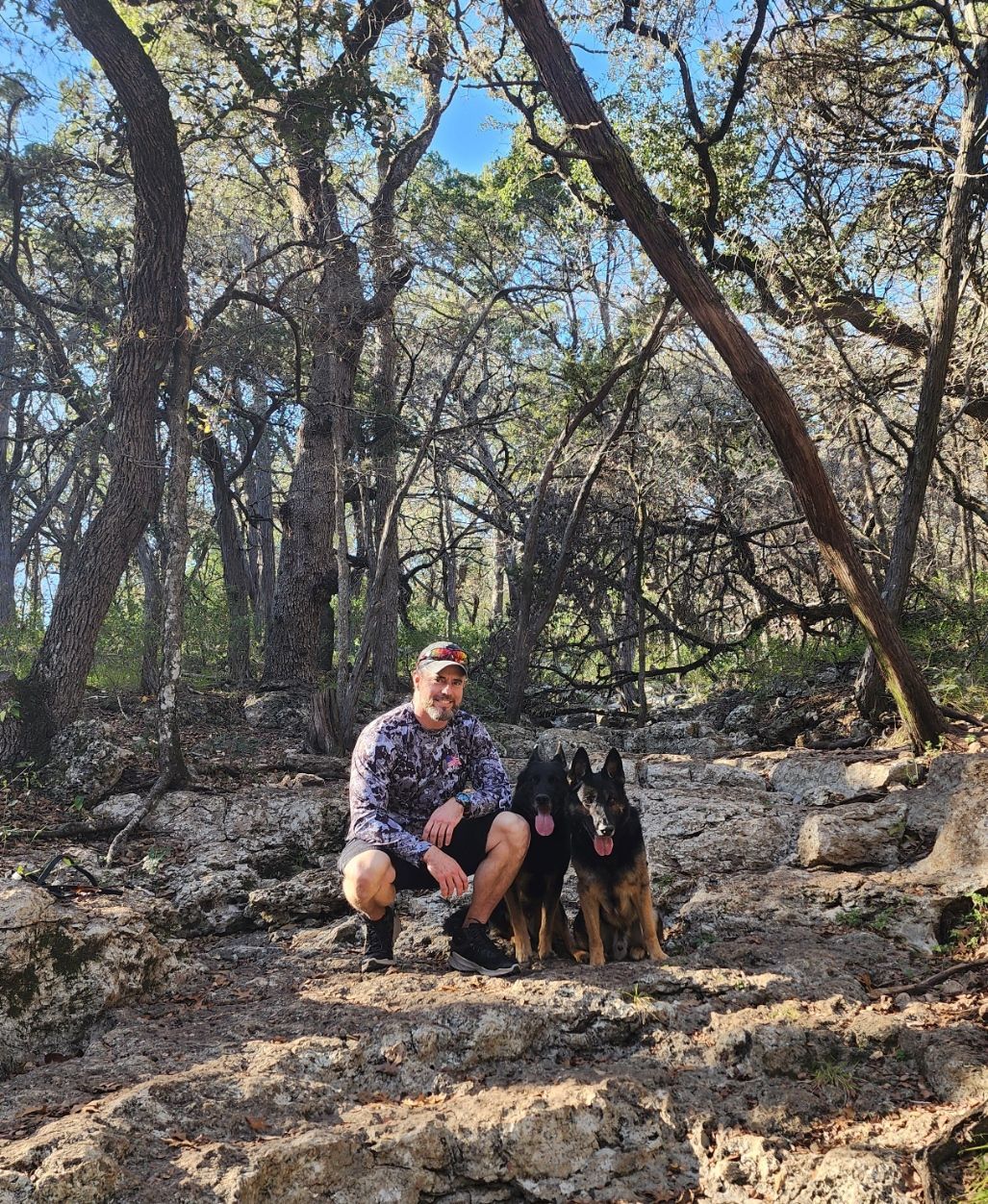 Man squats with two German Shepherds in a wooded, rocky area under a bright sky.