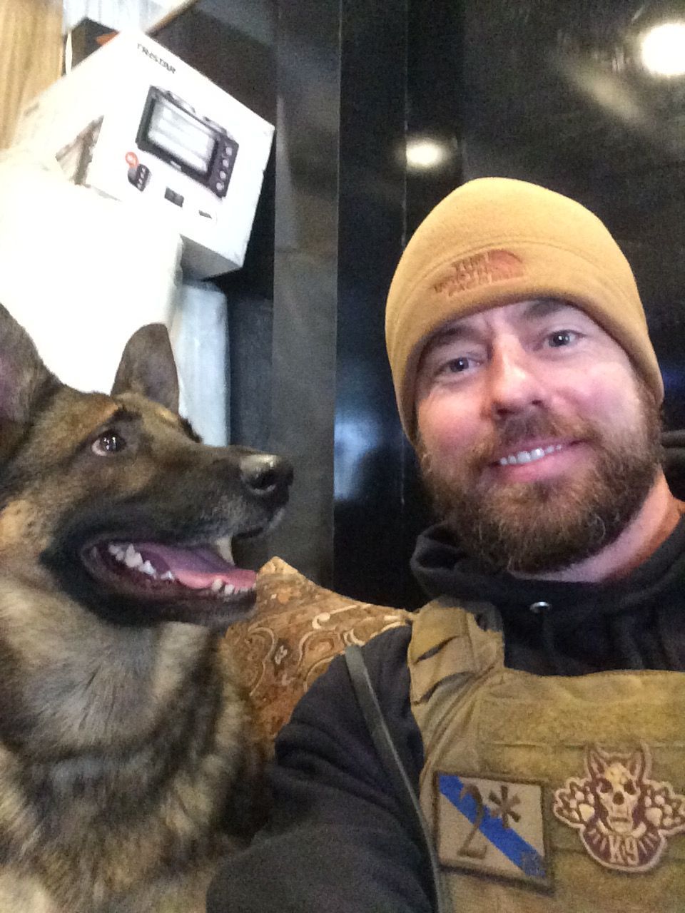 Man in tan beanie and tactical vest with a German Shepherd, posing for a selfie indoors.