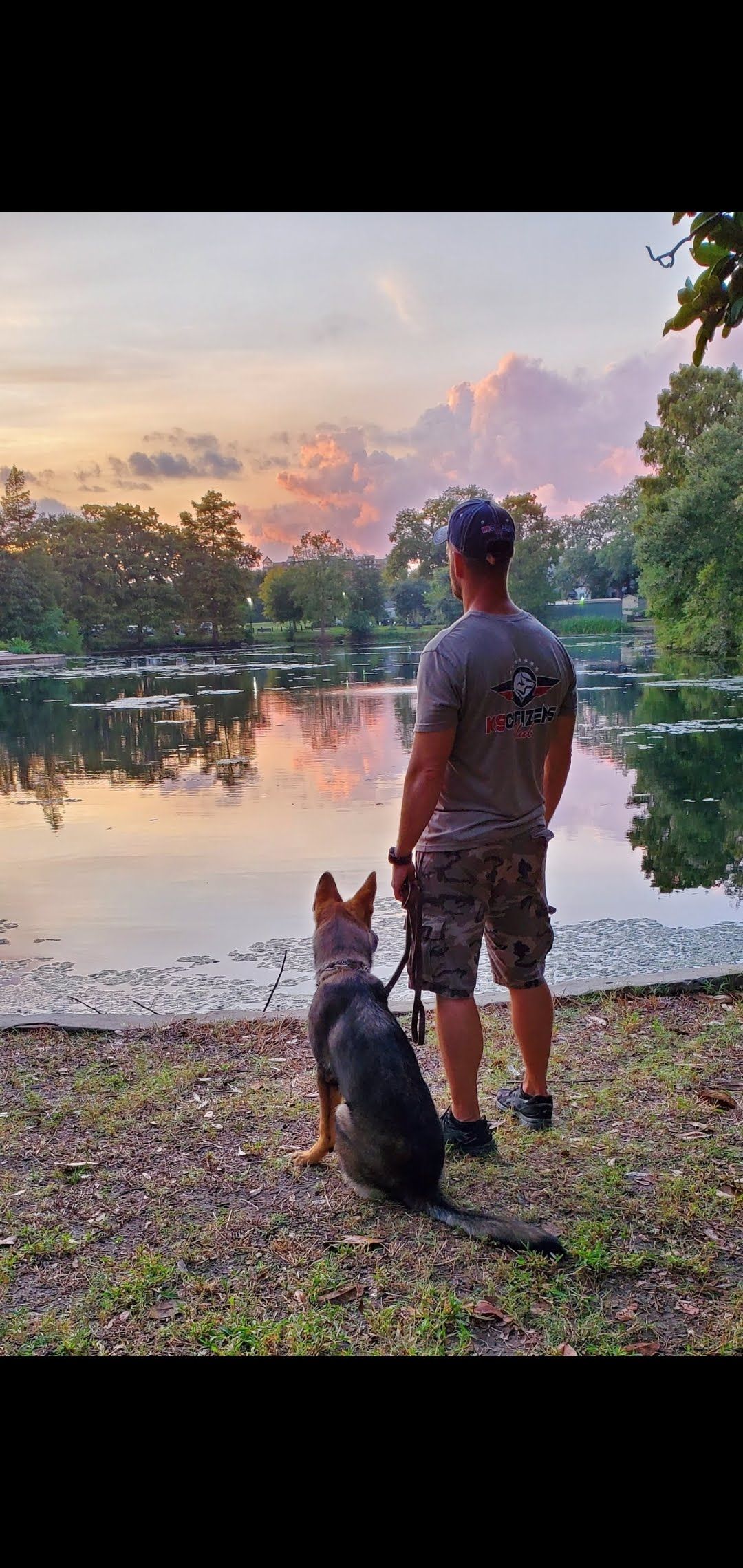 Man and dog gazing at a calm lake at dusk; trees and colorful sky are reflected in the water.