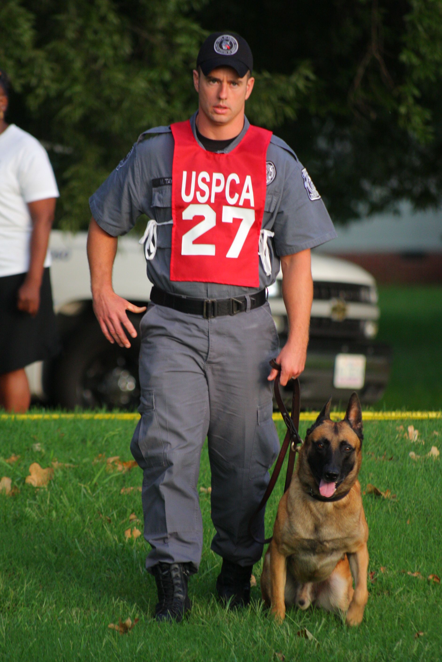 USPCA officer with a dog, wearing red vest. They stand on grass, the dog sitting.