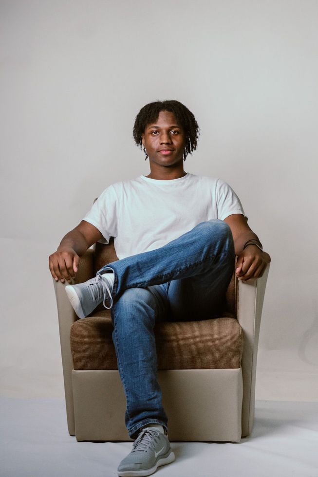 Young man sitting in a chair, legs crossed, wearing white tee, jeans, and sneakers; smiling at the camera.