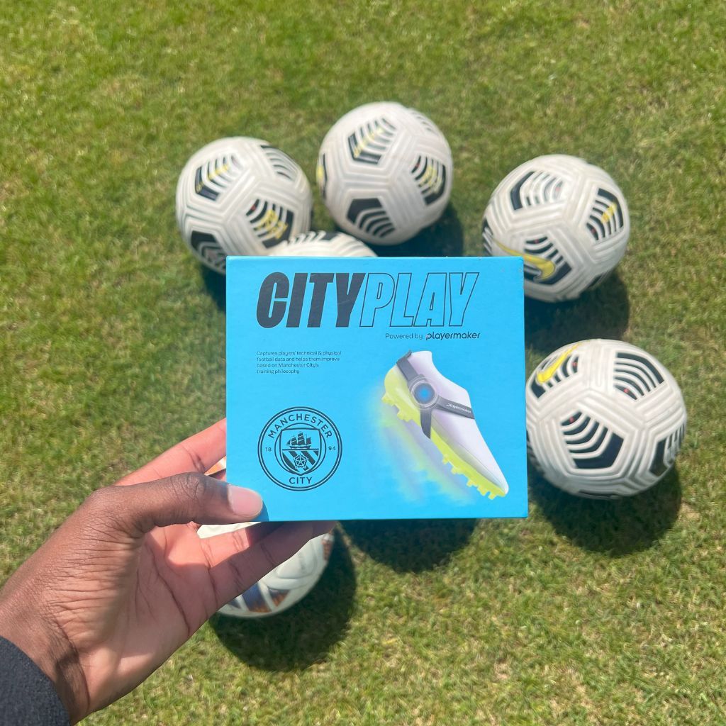A hand holds a turquoise box with a shoe graphic, surrounded by soccer balls on grass.