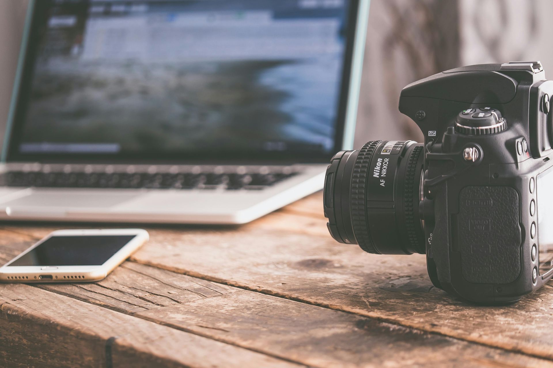 Camera, laptop, and phone on a wooden desk. The camera is facing right, and the laptop screen displays a blurry image.