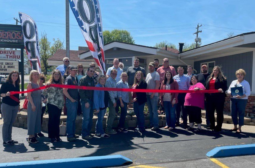 A group of people are standing in front of a building holding a red ribbon.