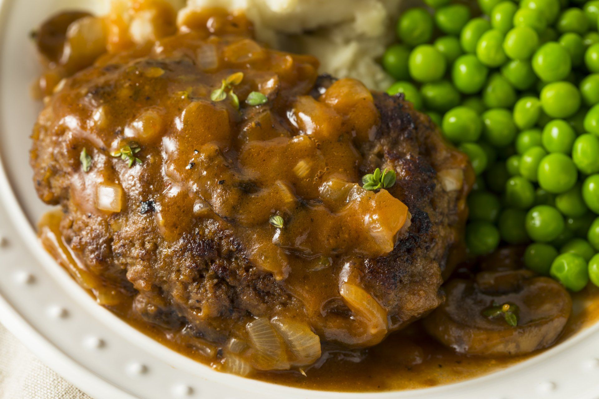 A white plate topped with a hamburger steak , mashed potatoes , peas and gravy.