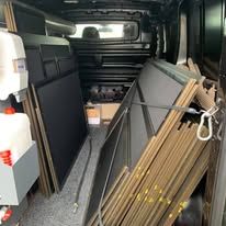 Inside a van cargo area, large black panels are stacked vertically on the left and right sides, secured by a strap.