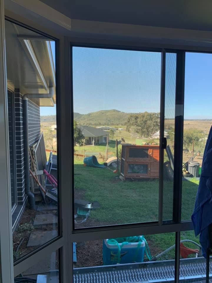 A view through a window overlooking a grassy yard with a small wooden enclosure, scattered items, and rolling hills beyond.