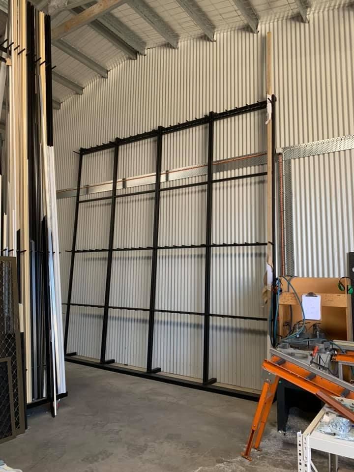 A tall, black metal storage rack for vertical sheet materials stands against a corrugated metal wall in a workshop.