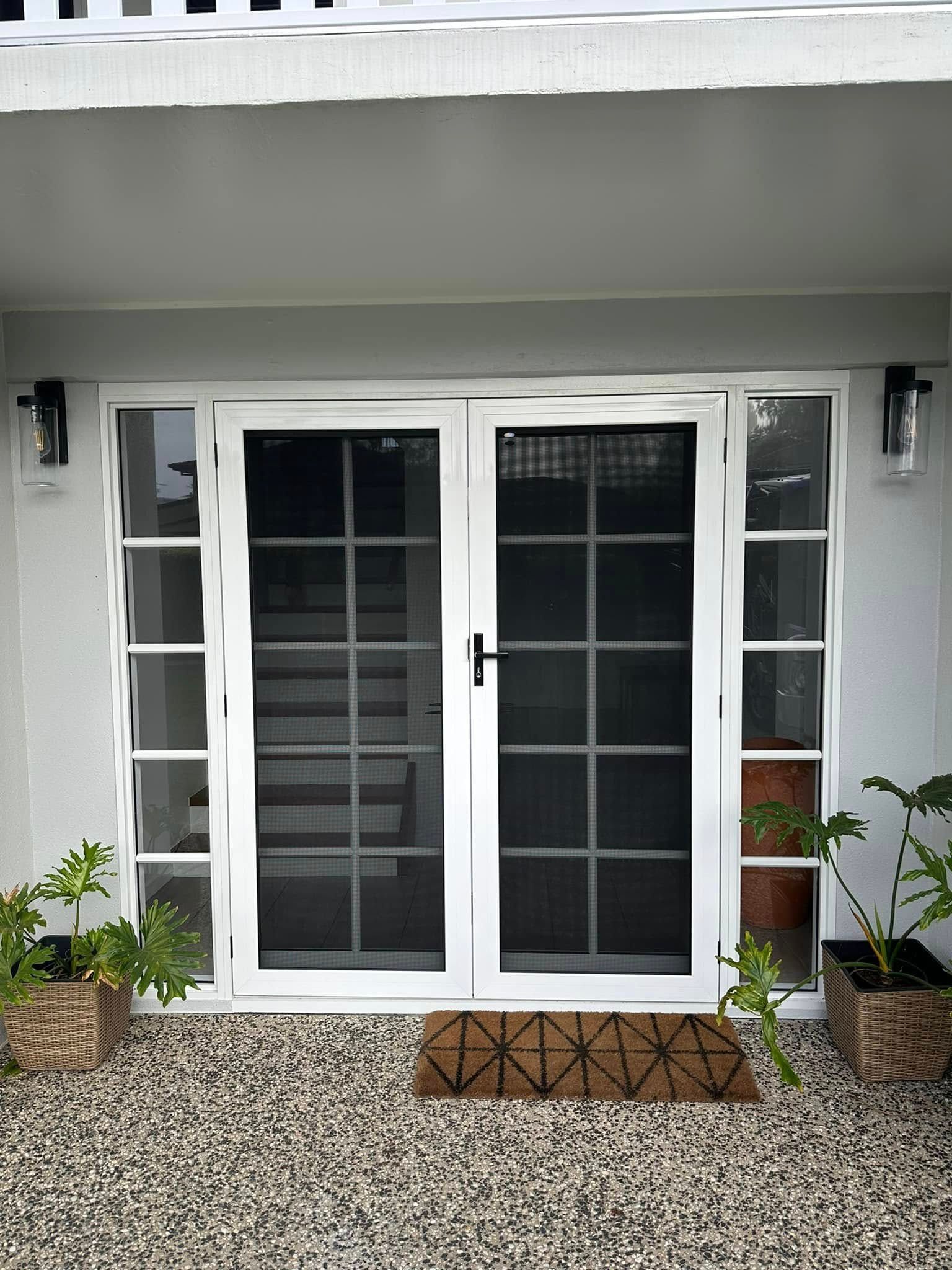 White double French doors with black screen inserts and matching side windows, framed by two potted plants on gravel.