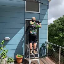 A person on a step ladder installs a screen door on the light blue exterior wall of a home near a balcony.