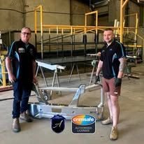 Two people in black work uniforms stand in a workshop behind a metal vehicle bumper, with logos at the bottom.