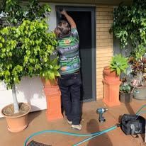 A person wearing a patterned shirt works on installing a screen door in a patio area with potted plants.