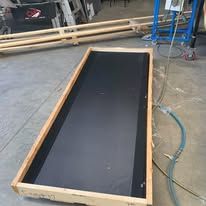 A rectangular wooden frame containing a large, flat black surface, situated on a concrete floor in a workshop.
