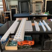 Several bundles of white metal window screen frames arranged on a dark workshop table, with a tool case underneath.