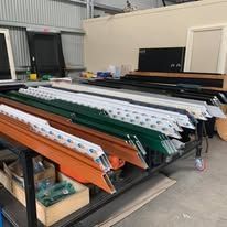 A workshop table holds organized rows of aluminum window frames in orange, green, black, and white colors.