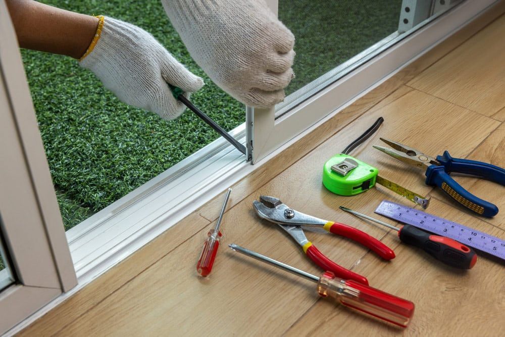 Gloved hands use a screwdriver on a sliding door frame, with various tools laid out on a wooden floor.