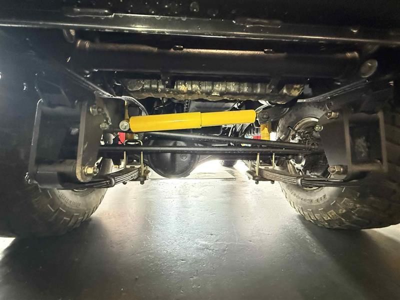 Underside view of a vehicle front suspension showing leaf springs, axle, steering stabiliser with yellow shock absorber, and off-road tyres. — Triple C Workshop in Springwood, QLD