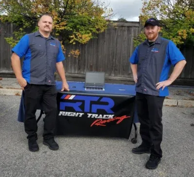 Two people in matching blue and gray company uniforms stand beside a table with a Right Track Racing logo outdoors.