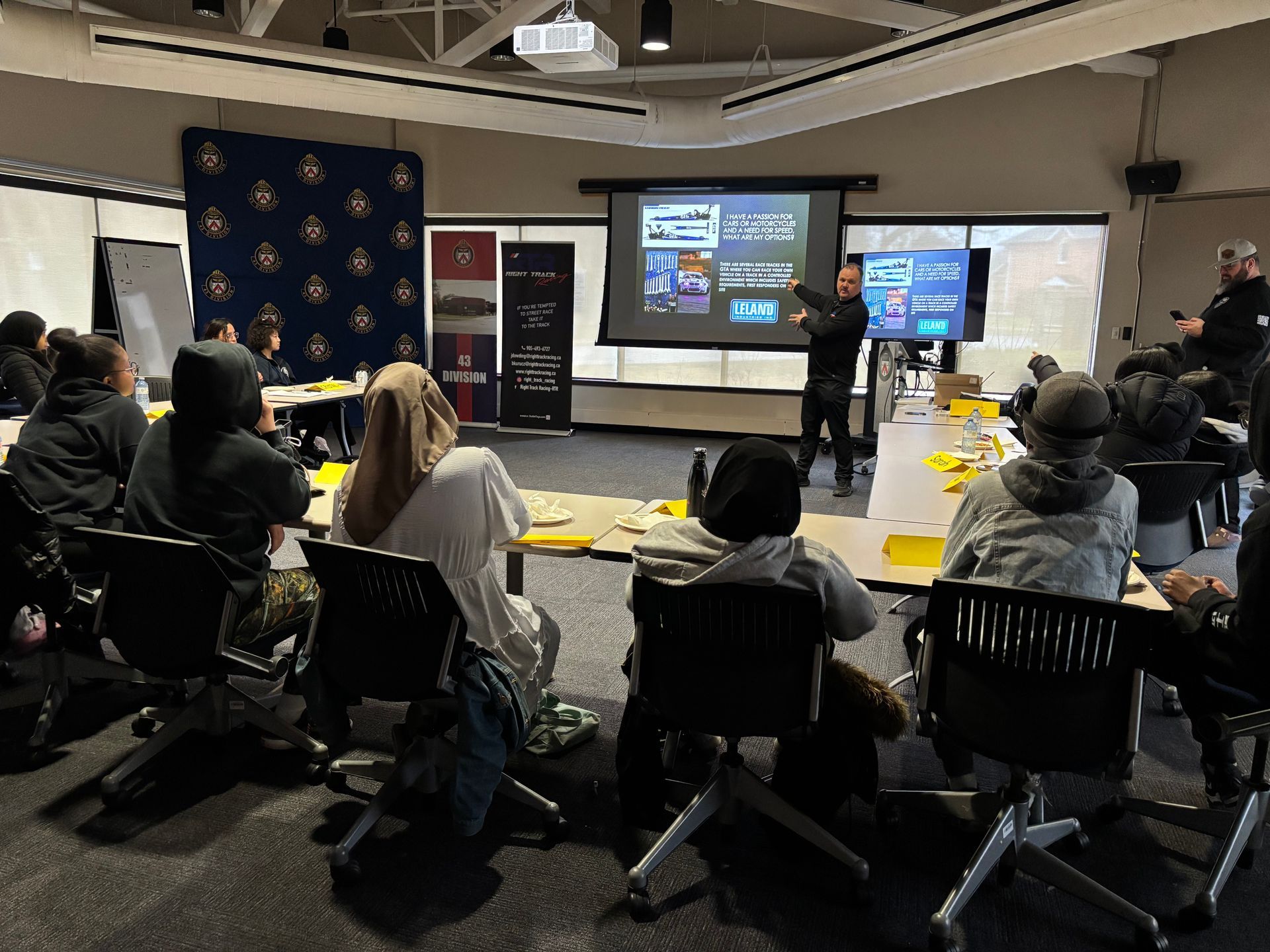 A speaker presents to a small audience in a room with a projector screen, desks, and chairs in a classroom setting.