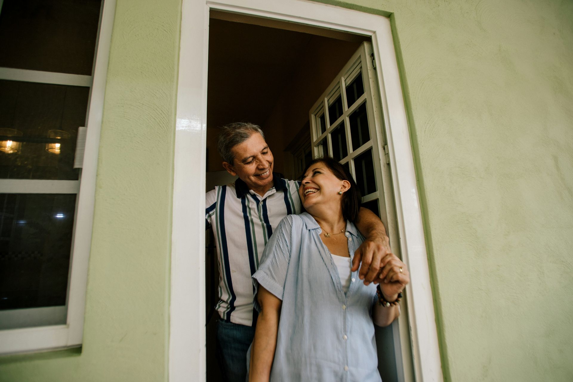 A smiling couple embraces in a doorway of a house with light green walls.