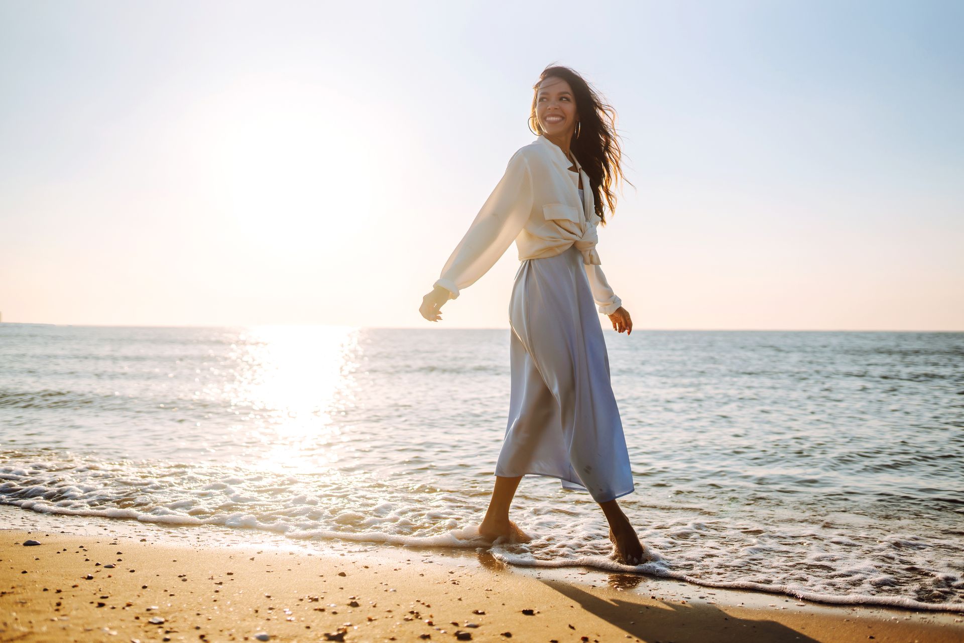 A person with long hair walks along the shoreline at sunset, smiling over their shoulder, wearing a light shirt and skirt.
