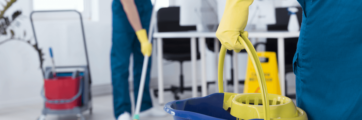 Cleaning crew in office. Individual wearing a glove holding a bucket in focus. Individual mopping in background blurred.