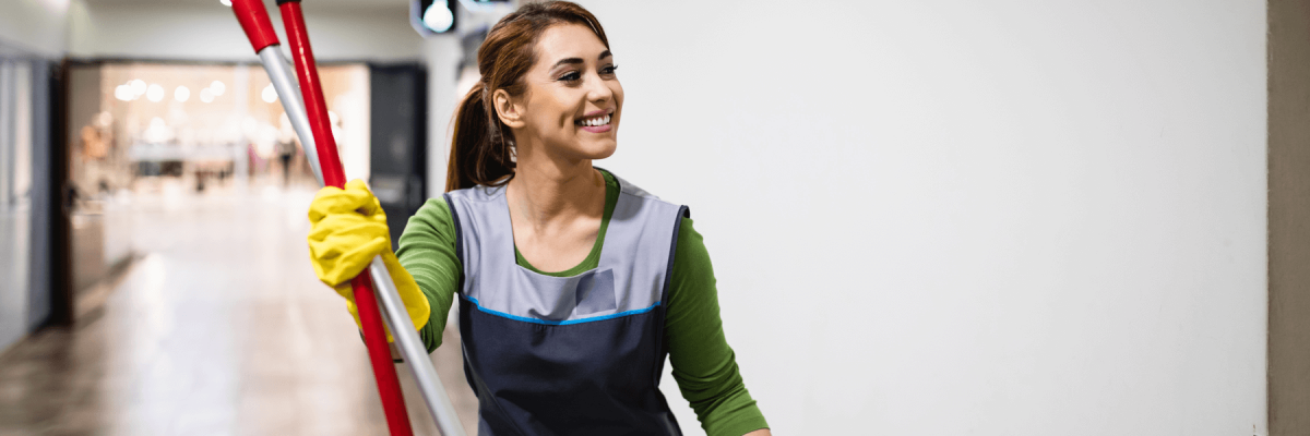 Smiling woman wearing cleaning gloves and apron carrying cleaning tools in a long hallway