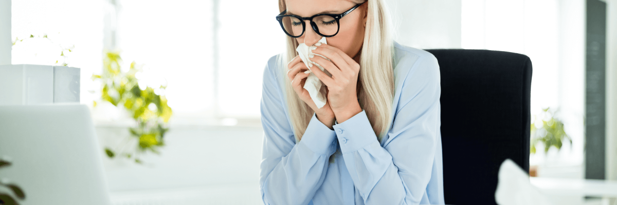 Blonde business woman wearing glasses sitting at office desk blowing nose