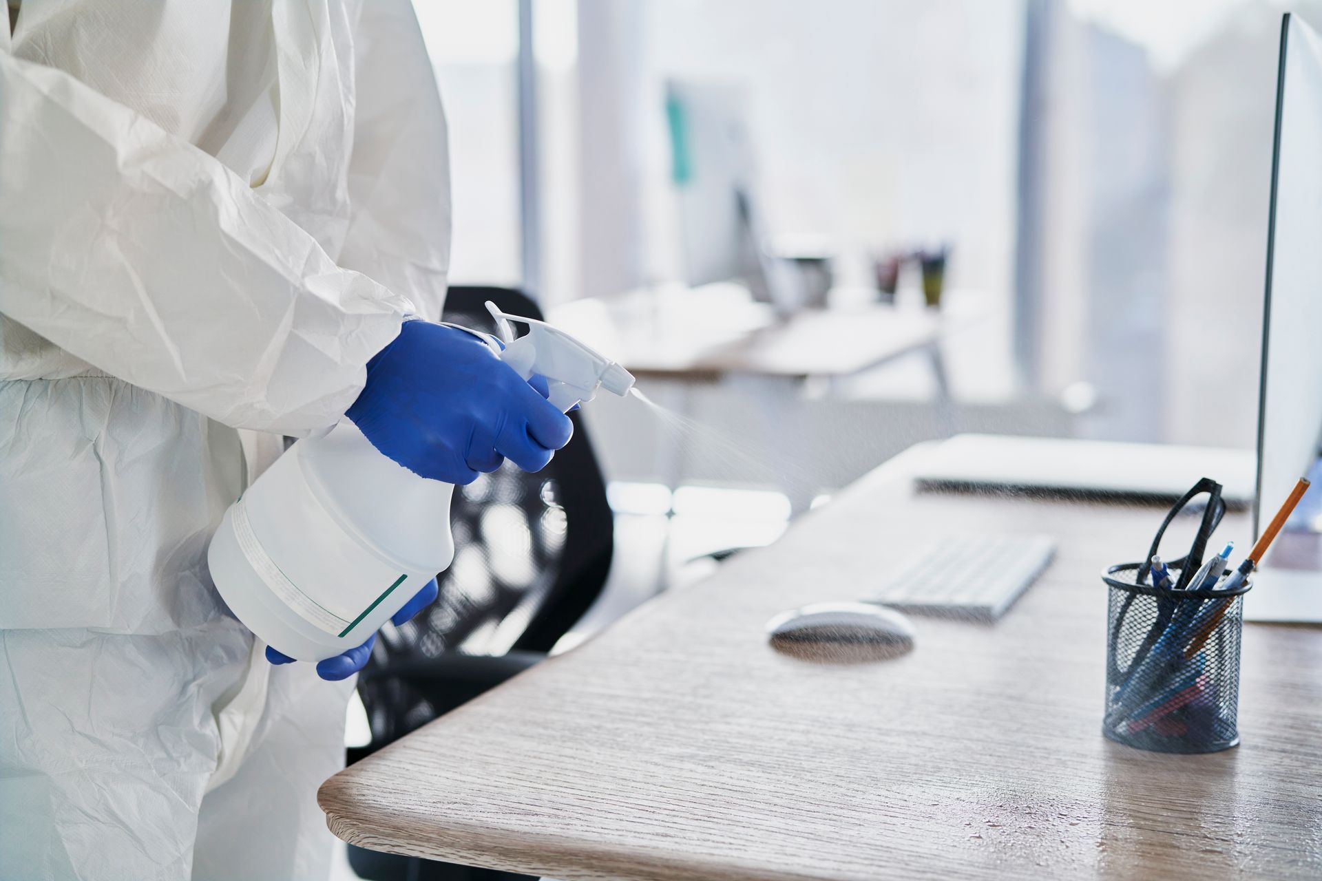 A person in a protective suit is disinfecting an office desk with a spray bottle.
