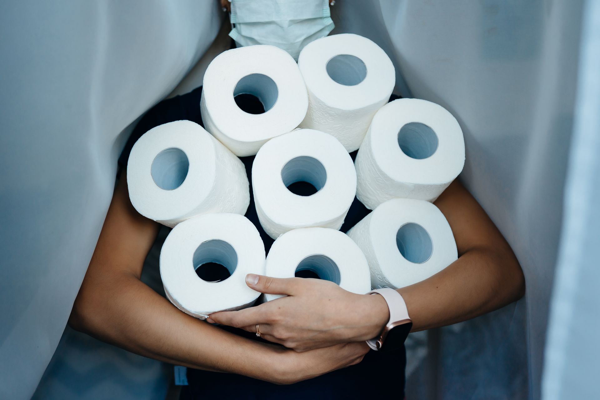 A woman wearing a mask is holding a pile of toilet paper rolls.