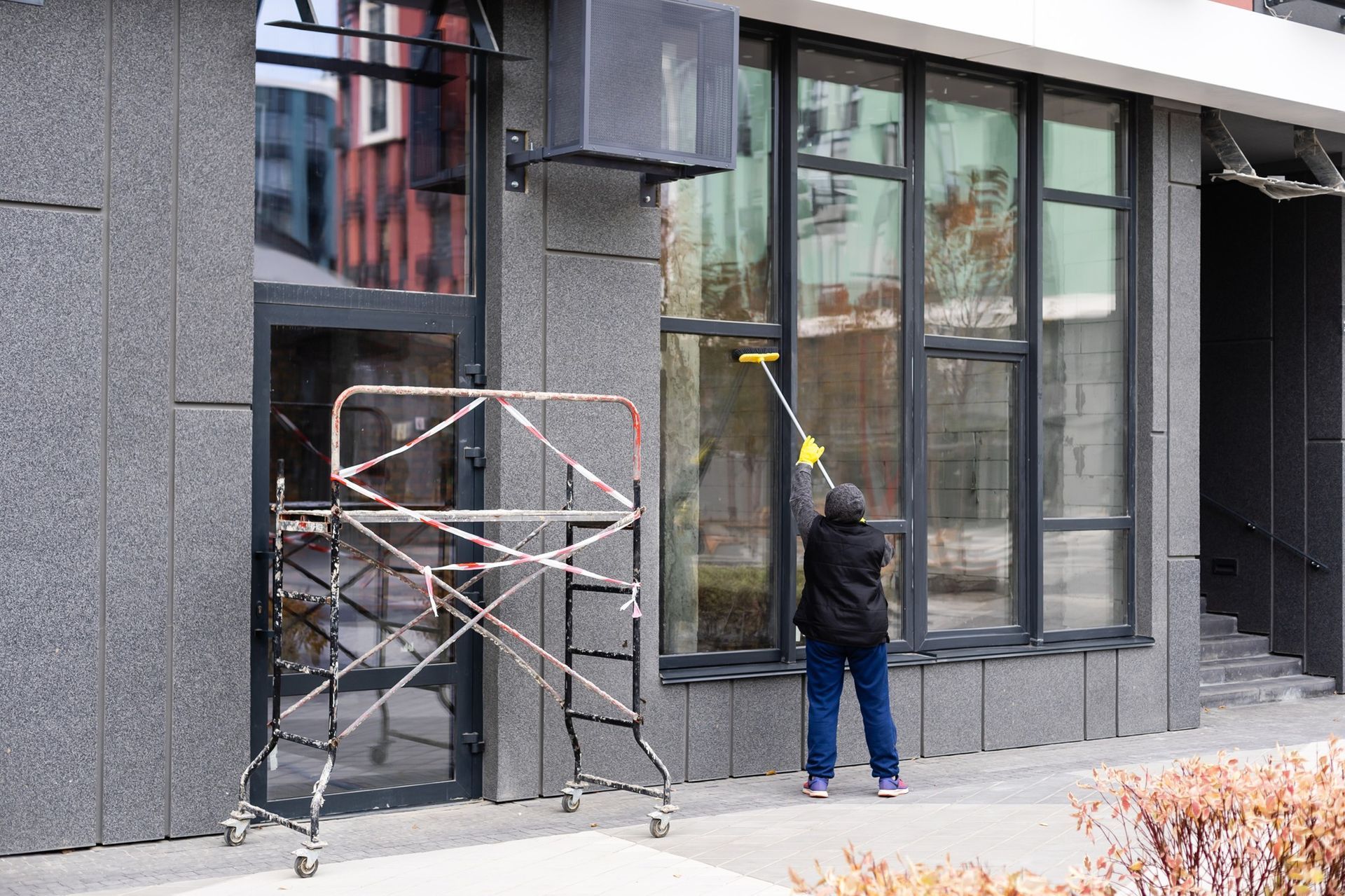 Person cleaning large window on a building, using a squeegee. Scaffolding stands nearby.