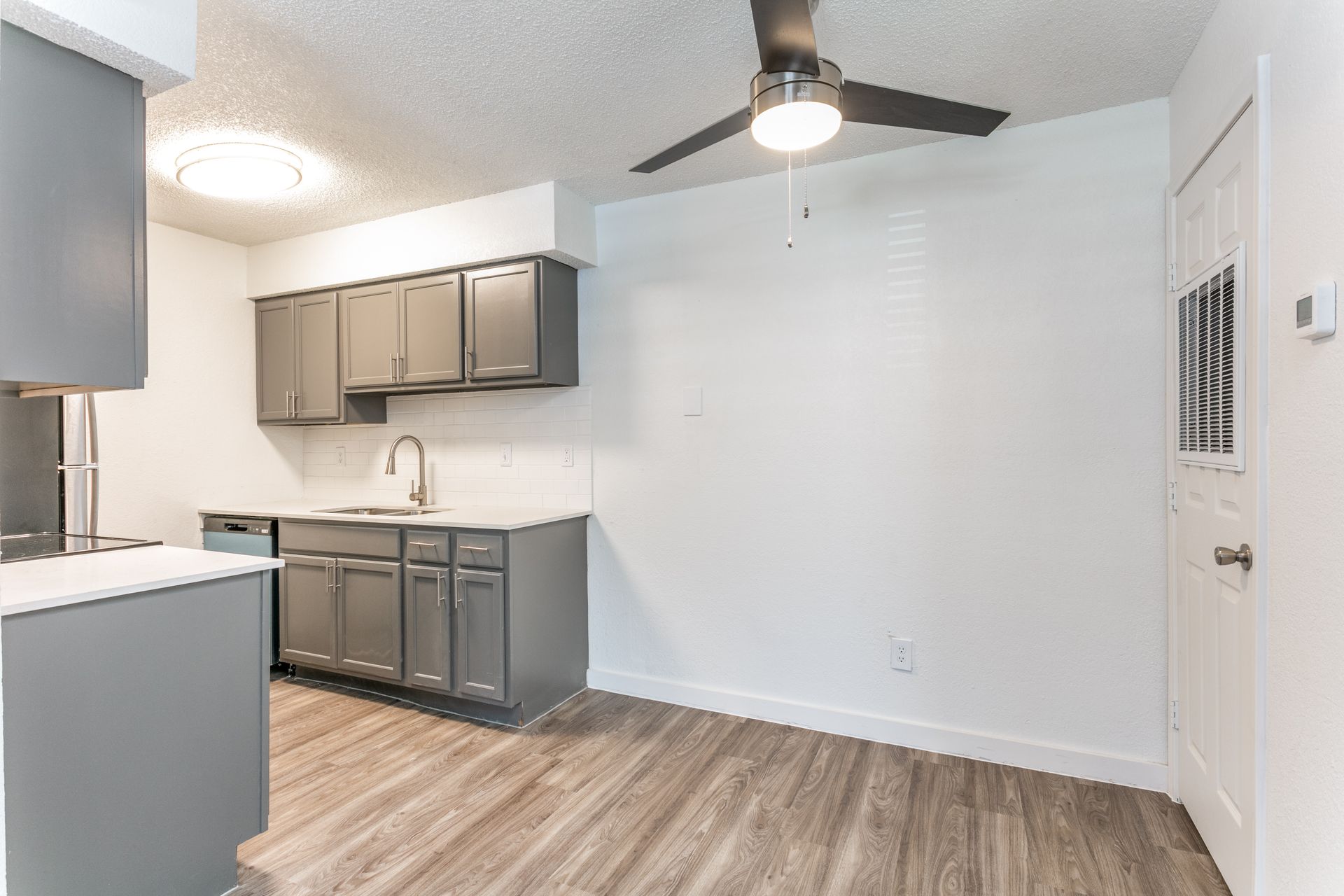 Grey kitchen with cabinets, sink, and a ceiling fan. Wooden floors and white walls.