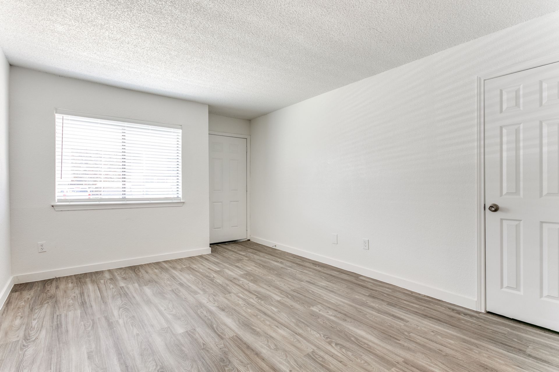 Empty, bright room with gray wood-look floor, window with blinds, and white doors and walls.