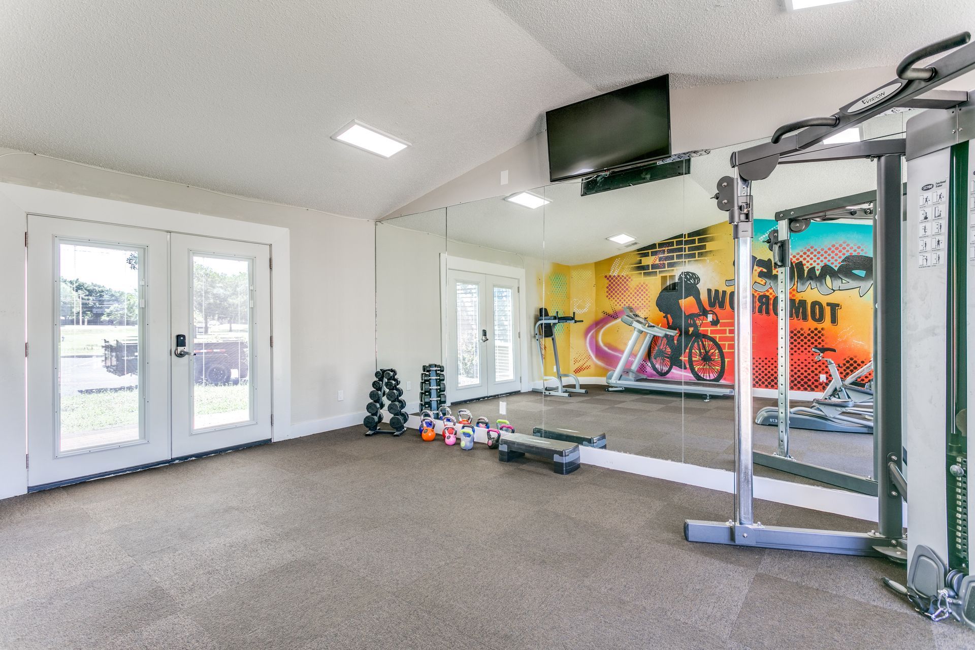Gym with cardio equipment, weights, and large mirror, with colorful wall mural and TV.