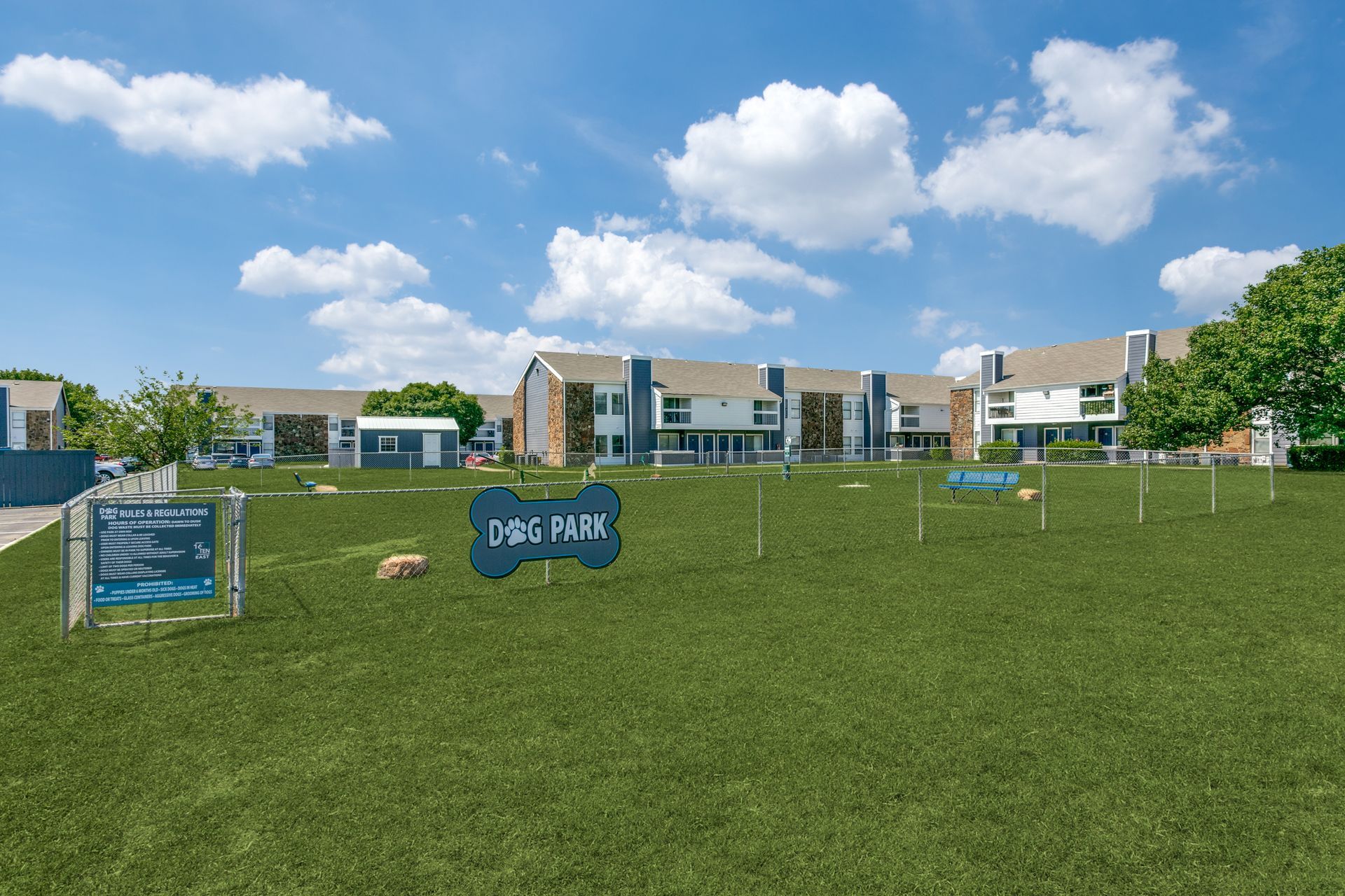 Dog park with sign, green grass, apartment buildings in background under blue sky.
