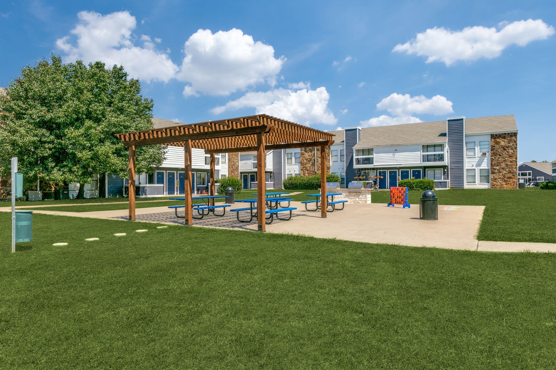 Park area with pergola, picnic tables, and playground equipment in front of apartment buildings, under a blue sky.