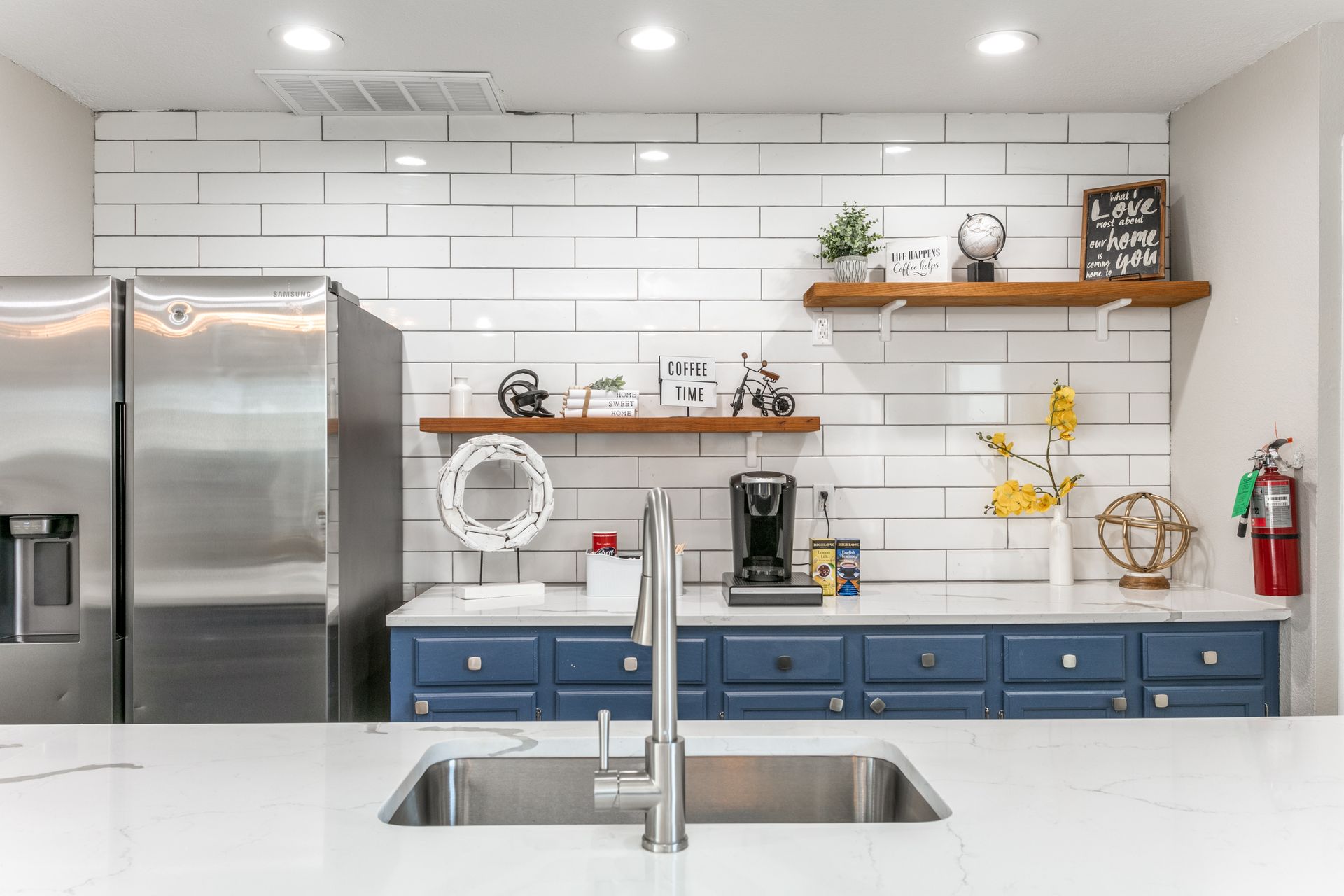 Modern kitchen with blue cabinets, white brick backsplash, stainless steel appliances, and a central sink.