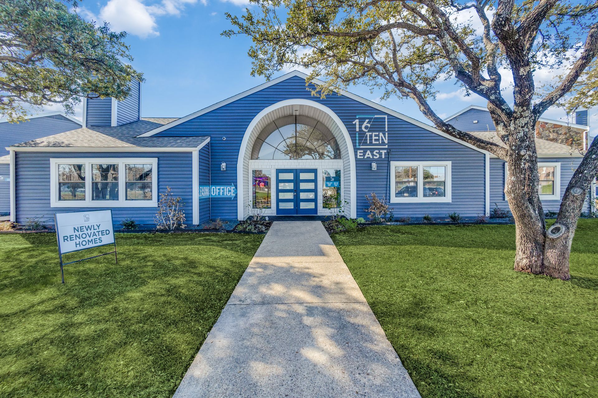 Blue building with arched entryway and a pathway, green lawn, sign on the left.