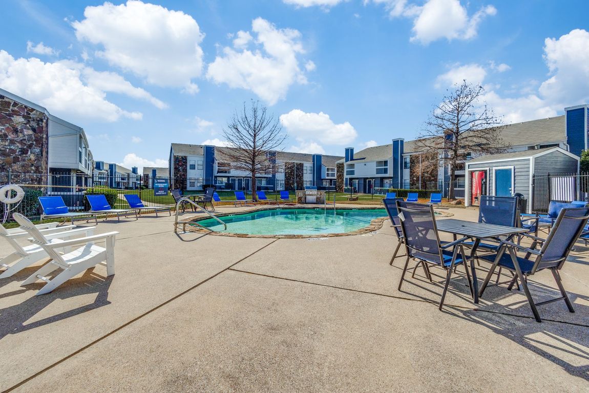 Swimming pool area with lounge chairs, tables, and apartment buildings on a sunny day.