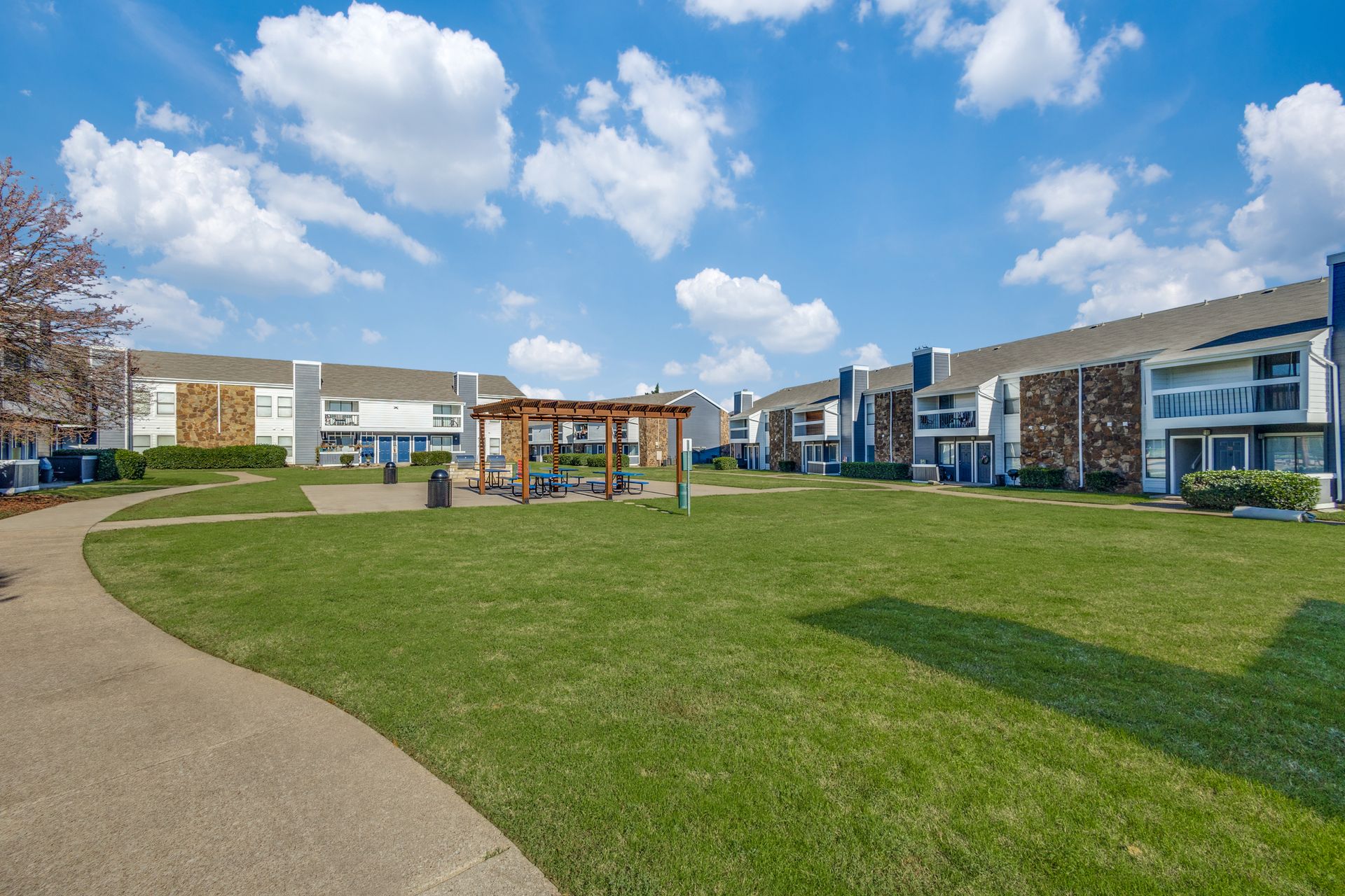 A grassy courtyard with a wooden pergola, surrounded by apartment buildings under a blue sky.
