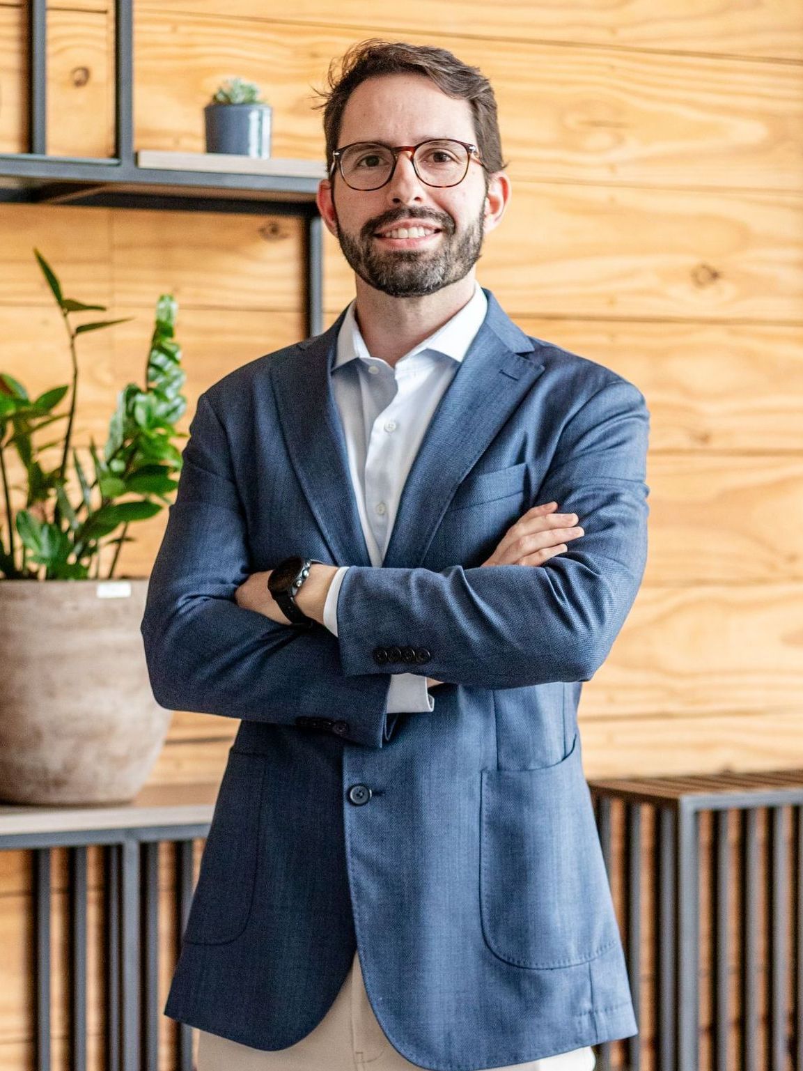 A man with a beard and glasses is standing with his arms crossed in front of a wooden wall.
