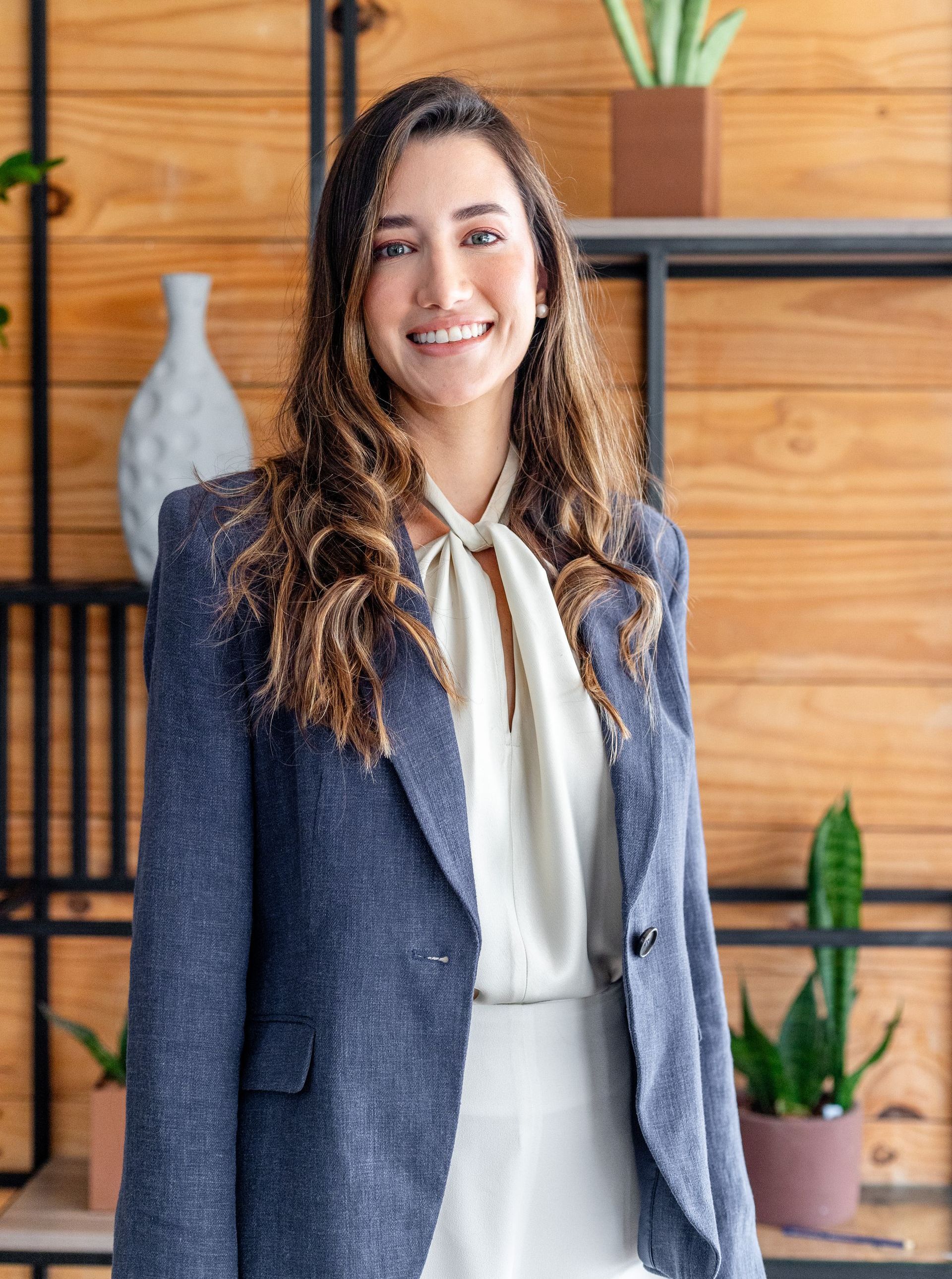 A woman in a blue jacket and white shirt is standing in front of a wooden wall.