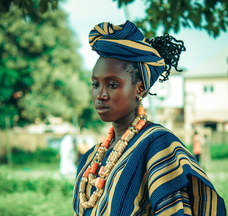 Woman in striped blue and yellow attire, wearing a headwrap and coral beads, outdoors.