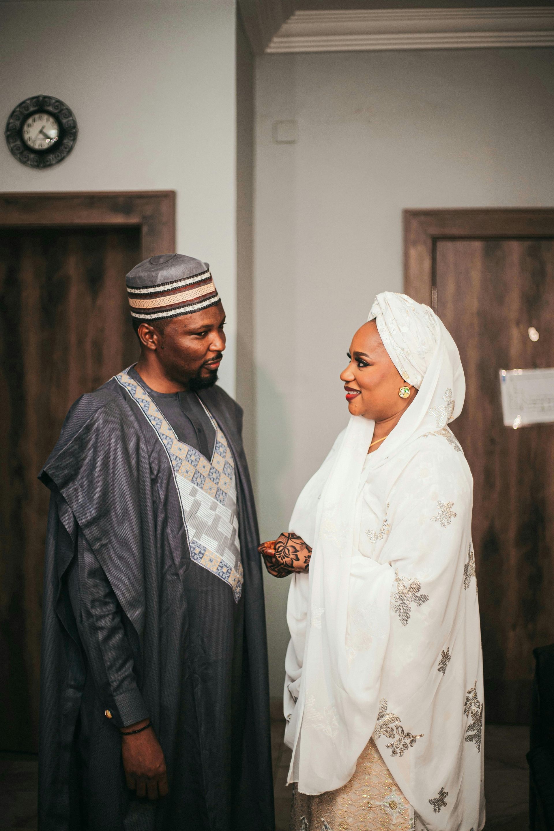 Man and woman in traditional attire face each other, smiling. Interior setting with doors and a clock on the wall.