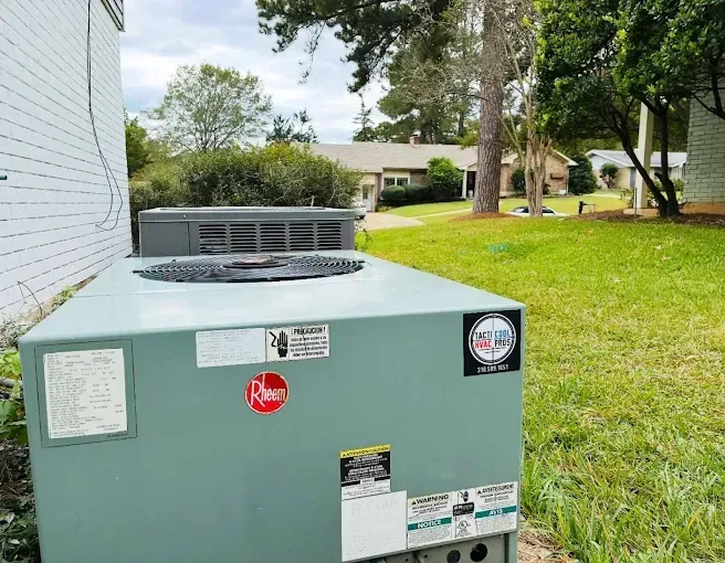Air conditioning unit on a grassy lawn next to a white building. Houses and trees in the background.