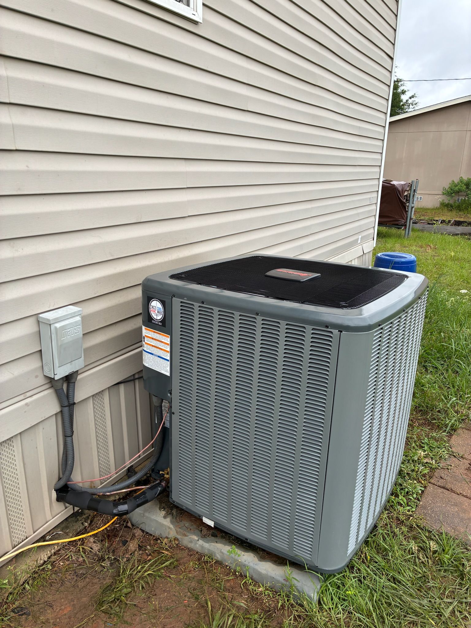 An air conditioning unit against a light brown siding wall with an electrical box.