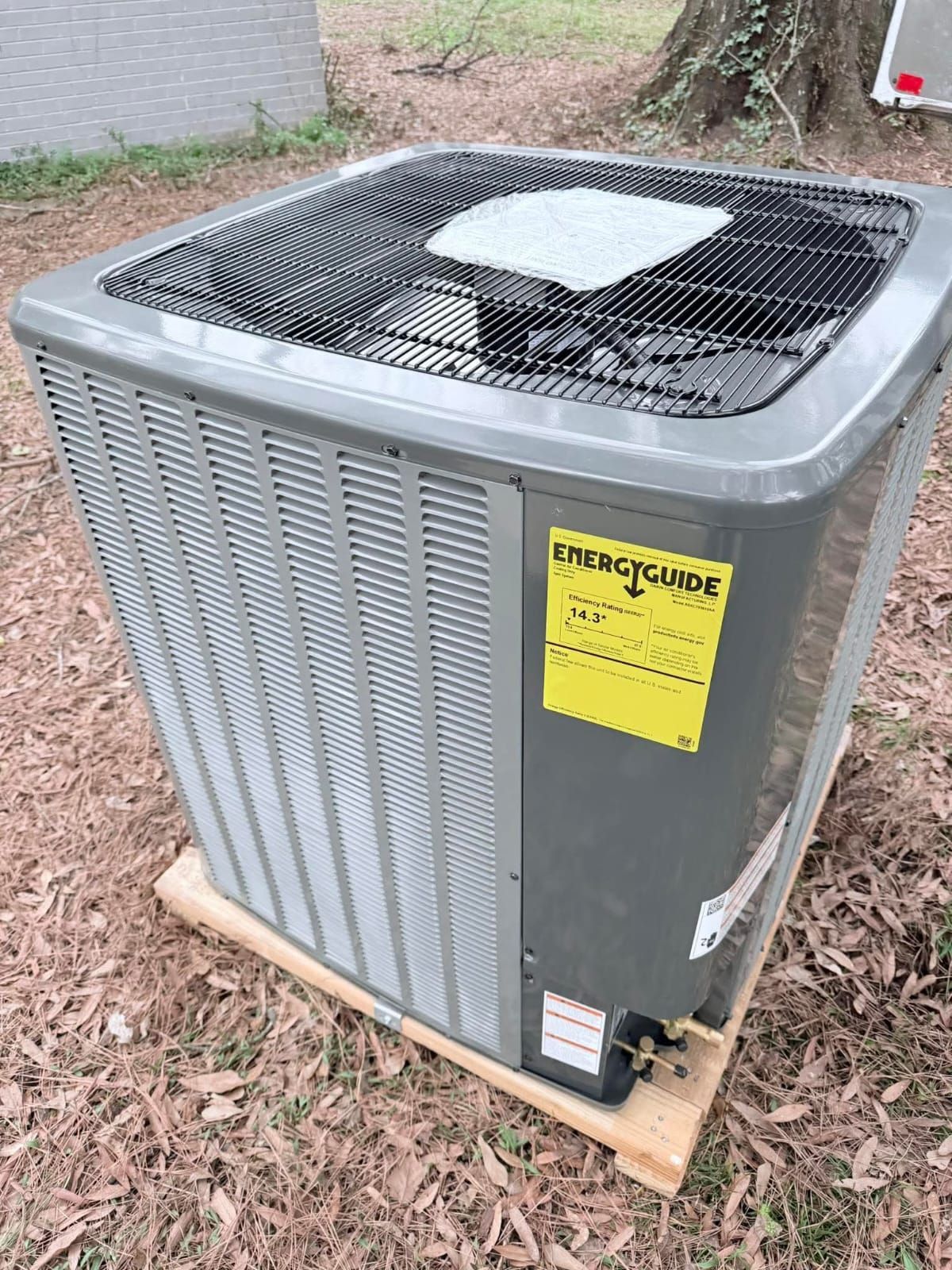 Gray air conditioning unit on a wooden pallet outdoors, with a yellow energy guide label.